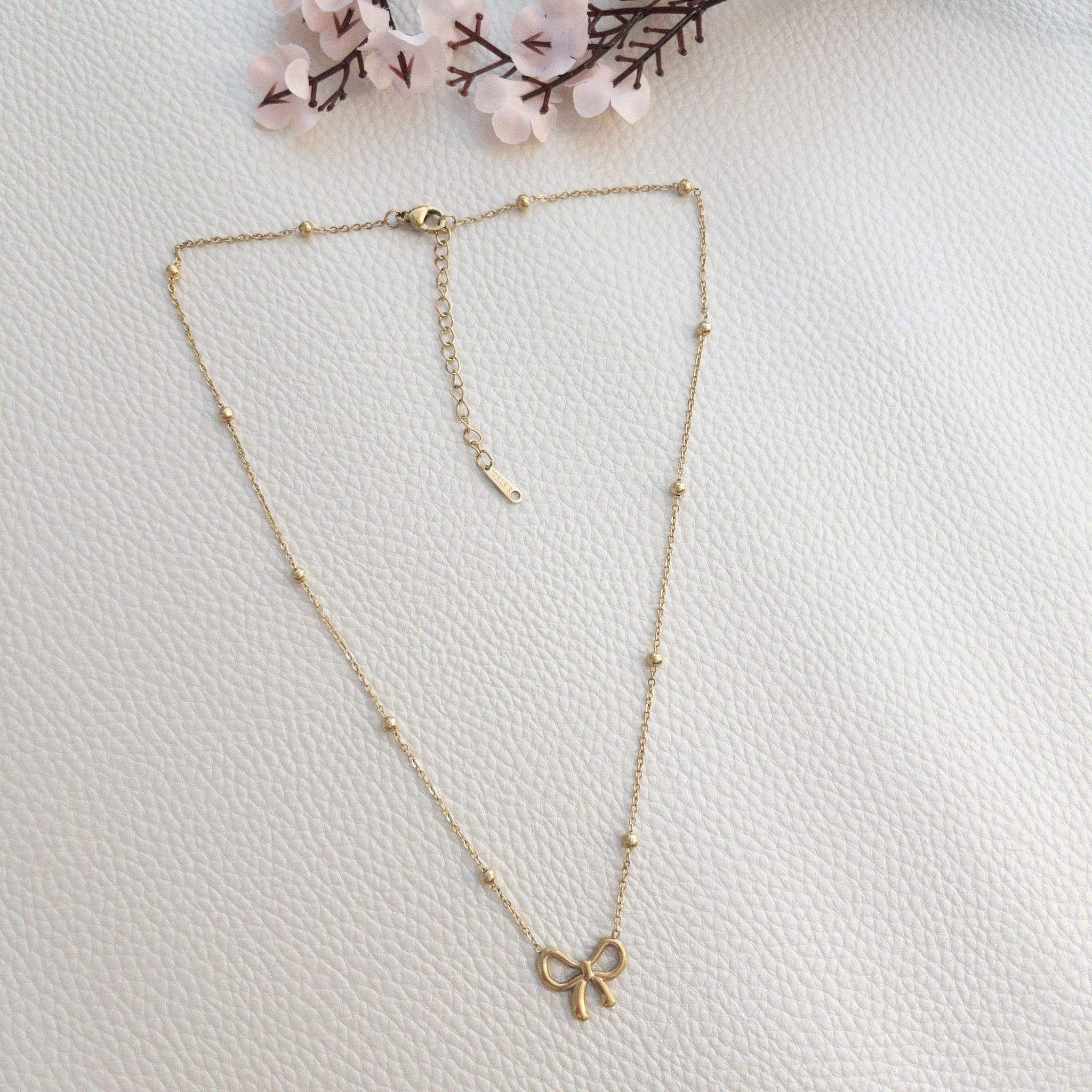 Collar Lazo (2) Collar dorado con lazo sobre fondo blanco texturizado y flores.